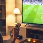 Person holding a remote control and watching a free World Cup stream on a flat-screen TV in a cozy living room with candles and shelves in the background.
