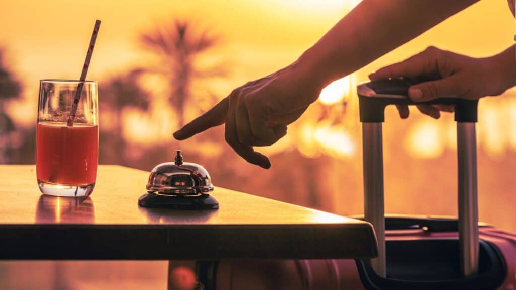 A hand presses a service bell on a table next to a glass of juice and luggage handle, with a warm sunset background&mdash;a moment hotel concierges wish guests ask for often, blending attentive service with relaxing ambiance.