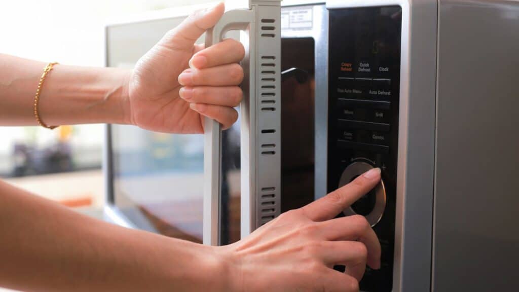 A person opens a microwave oven door with one hand while pressing a button on the control panel with the other hand.