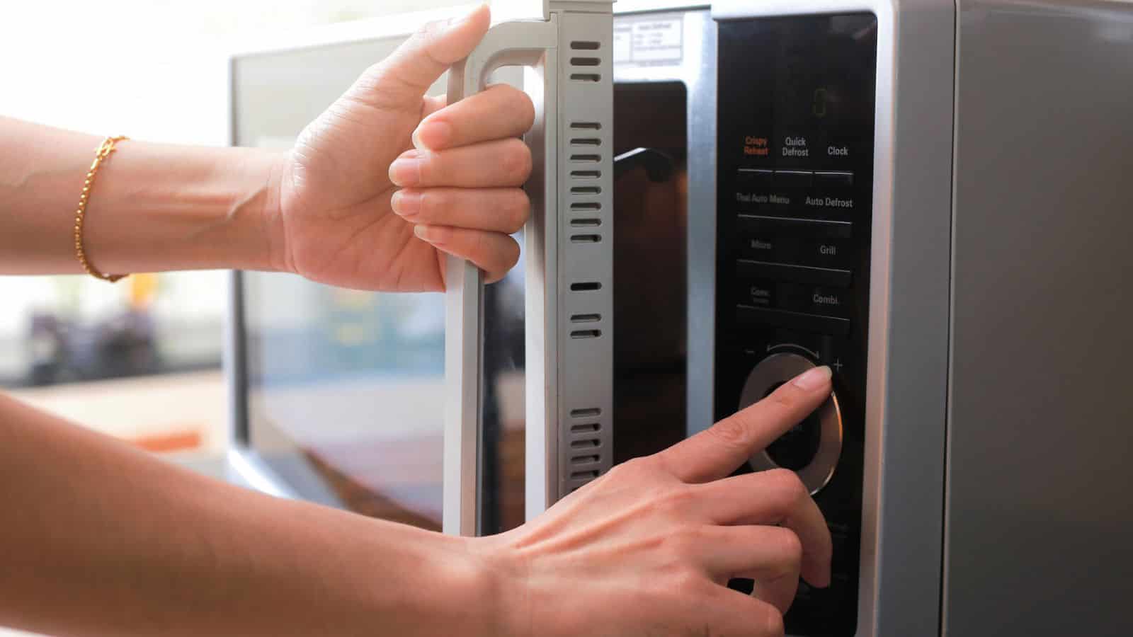 A person opens a microwave oven door with one hand while pressing a button on the control panel with the other hand.