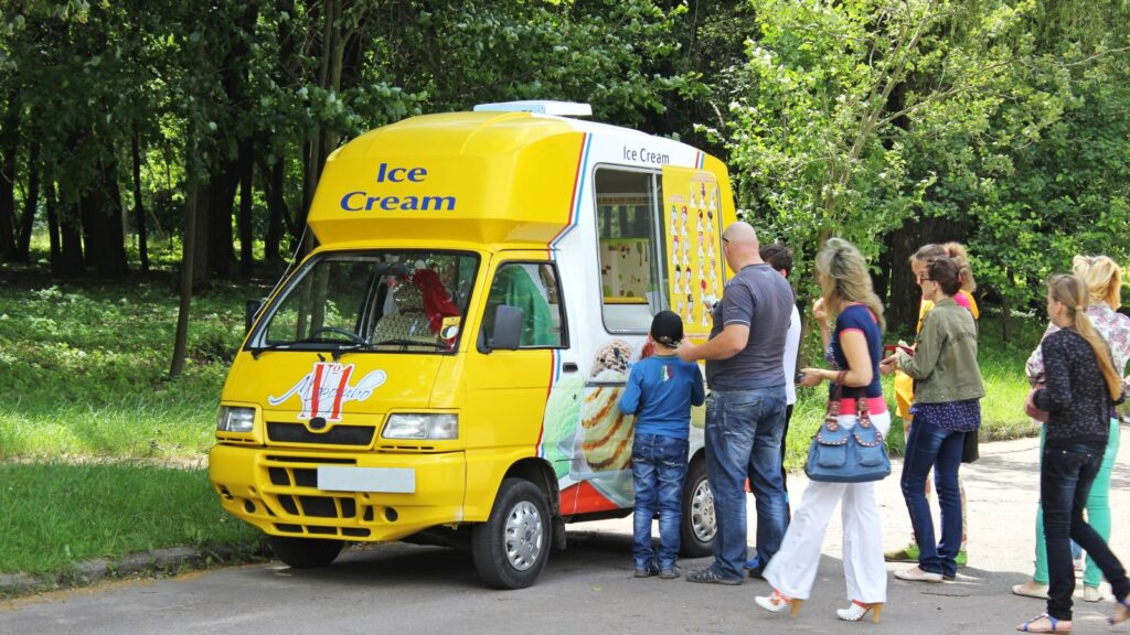 A group of people, including children, stand in line at a yellow ice cream truck parked on a tree-lined street, eagerly waiting for their favorite ice cream truck treats.