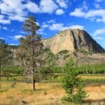 Grassland with scattered trees in the foreground, a river, and a rocky mountain rise under a blue sky with white clouds in the background.