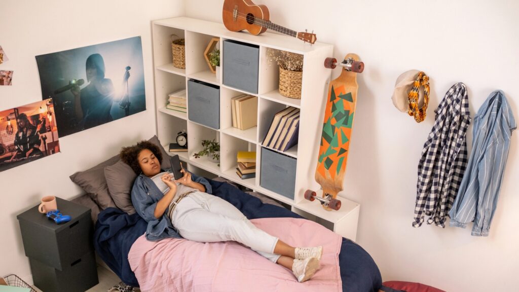 Young person lying on a bed in a tidy bedroom with a skateboard, musical instruments, and posters, looking at a smartphone.