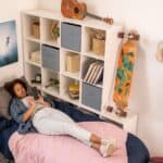 Young person lying on a bed in a tidy bedroom with a skateboard, musical instruments, and posters, looking at a smartphone.
