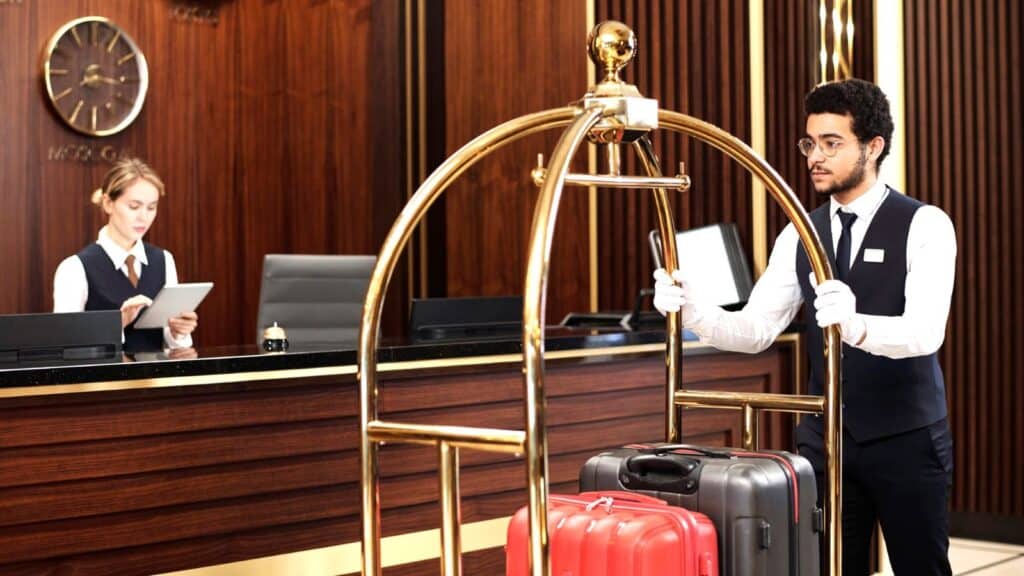 A hotel staff member pushes a luggage cart with suitcases while another employee, one of the attentive hotel concierges, works at the reception desk in a lobby, ready to assist with any requests guests may ask.