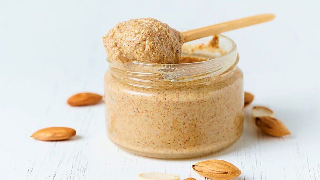 A glass jar of almond butter with a wooden spoon rests on a white surface, surrounded by whole almonds and almond shells.