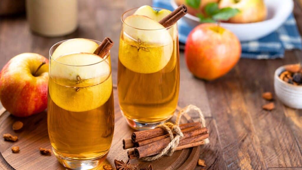 Two glasses of apple cider garnished with apple slices and cinnamon sticks on a wooden tray, surrounded by apples and spices.