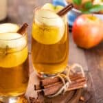 Two glasses of apple cider garnished with apple slices and cinnamon sticks on a wooden tray, surrounded by apples and spices.