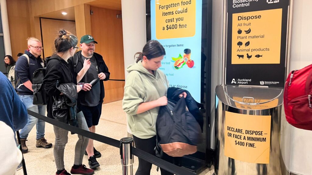 People stand in line at an airport near a bin labeled for declaring or disposing of prohibited food items, with signs warning of a minimum $400 fine for unreported goods and helpful information about which foods are allowed through airport security.