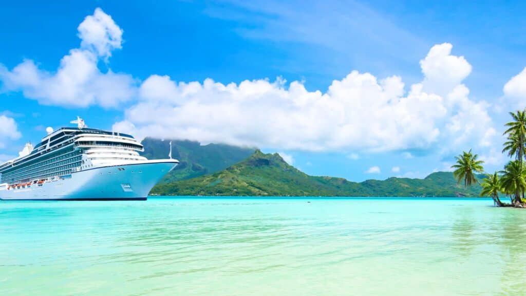 A large white cruise ship sails near a tropical island with green mountains, turquoise water, and palm trees under a blue sky with clouds.