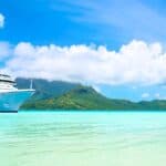 A large white cruise ship sails near a tropical island with green mountains, turquoise water, and palm trees under a blue sky with clouds.