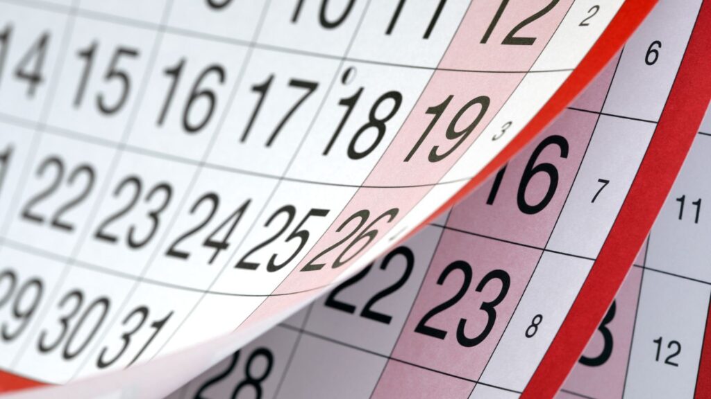 Close-up of overlapping pages from a paper calendar showing dates, with a page being flipped to reveal the next month&rsquo;s dates underneath.