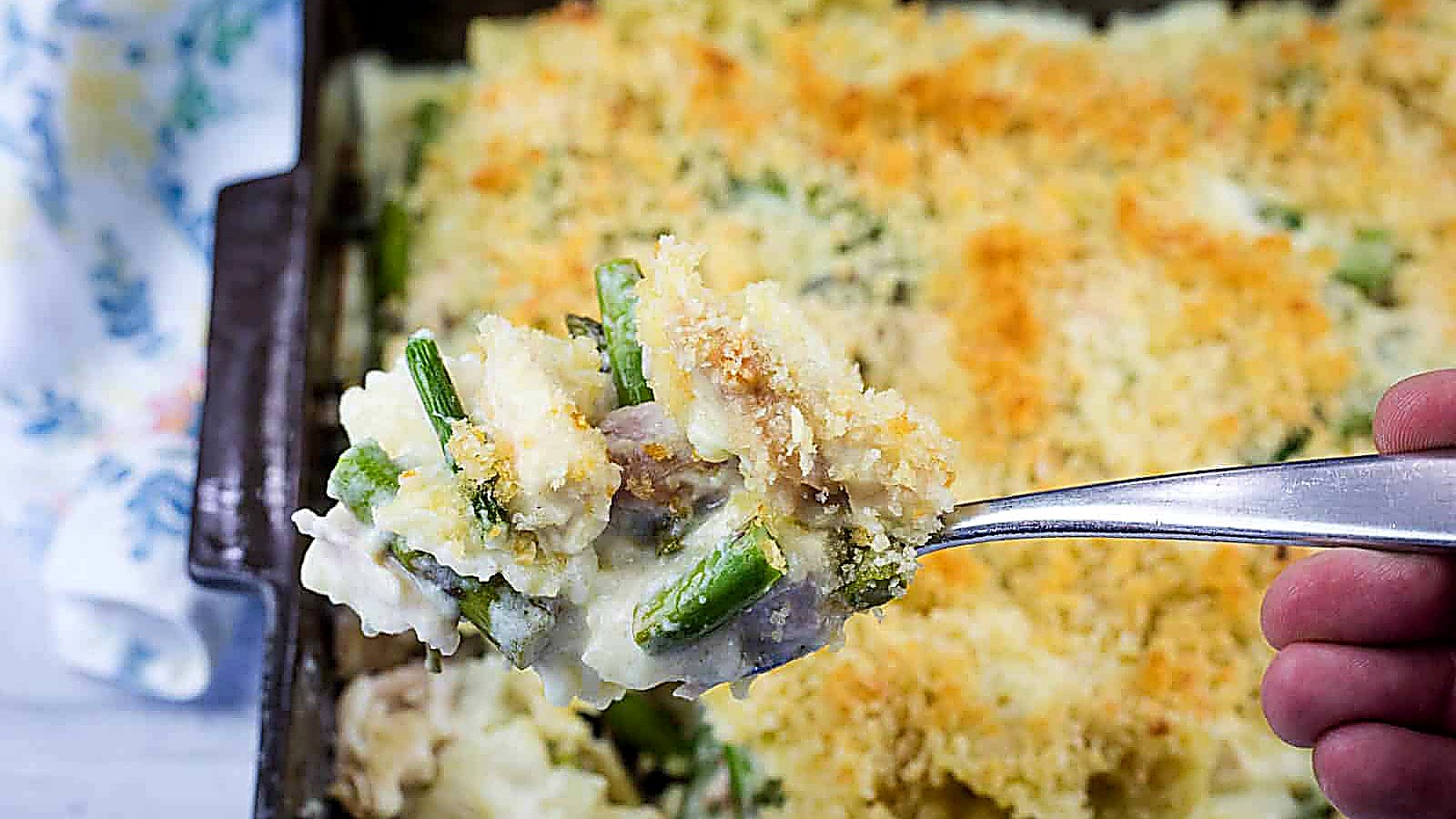 A hand holds a spoonful of Gruy&egrave;re Chicken Asparagus Pasta casserole topped with breadcrumbs over a baking dish filled with the same casserole.