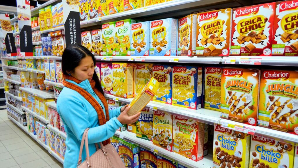 A woman in a blue coat reads a cereal box label while standing in front of shelves stocked with various breakfast cereals in a grocery store.
