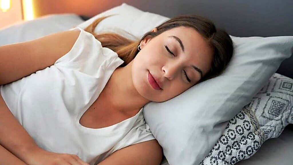A woman with long hair sleeps on her side in bed, resting her head on a pillow and wearing a white shirt, perhaps unwinding after learning about what is red light therapy and its relaxing benefits.