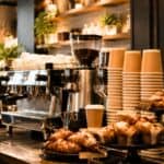 Coffee shop counter with stacked paper cups, pastries, a large espresso machine, and a menu board displaying drink prices. Shelves with plants and mugs decorate the background.