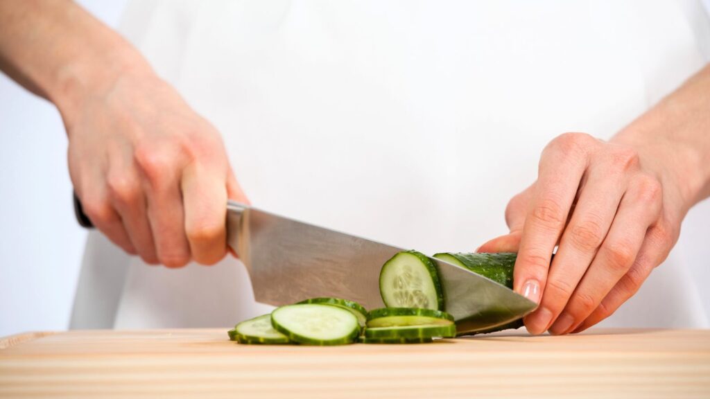 A person slices a cucumber on a wooden cutting board with a large kitchen knife, skillfully preparing ingredients for the World&rsquo;s Best Female Chef cucumber dessert.