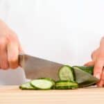 A person slices a cucumber on a wooden cutting board with a large kitchen knife, skillfully preparing ingredients for the World’s Best Female Chef cucumber dessert.