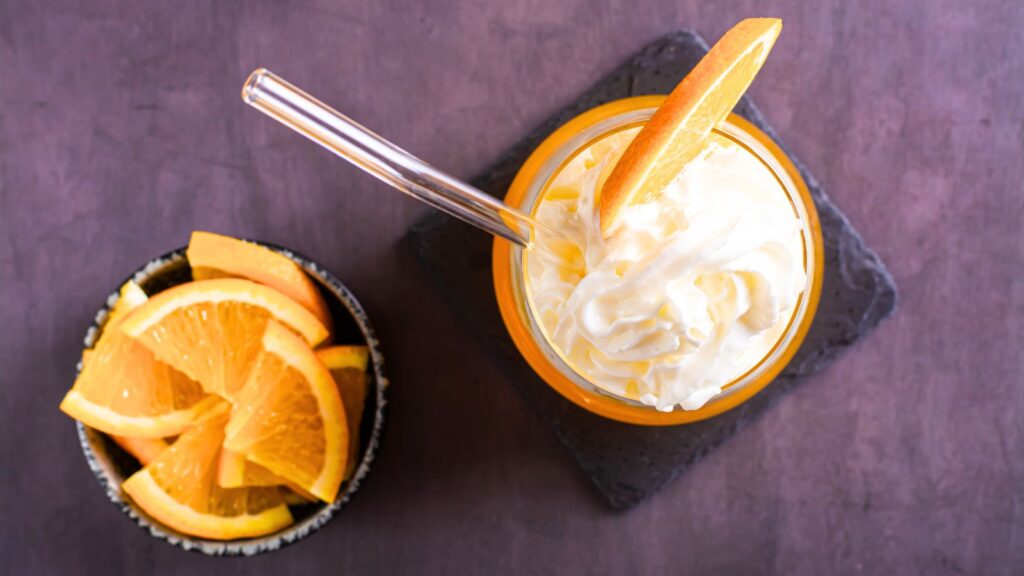 A glass of whipped cream-topped orange drink with a metal straw and orange slice beside a small bowl of orange wedges on a dark surface.