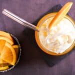 A glass of whipped cream-topped orange drink with a metal straw and orange slice beside a small bowl of orange wedges on a dark surface.