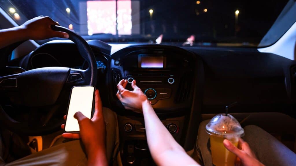 Two people sit in a car at night; one uses a phone, the other adjusts the radio. A drink is on the passenger’s lap and a projector screen is visible through the windshield.