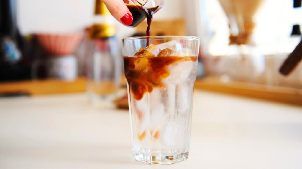 A hand pours dark liquid, likely coffee, into a clear glass filled with ice cubes on a light-colored surface.