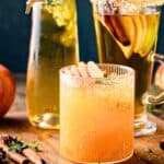 Three glasses of apple cider garnished with apple slices and spices, surrounded by fresh apples, cinnamon sticks, and star anise on a wooden board.