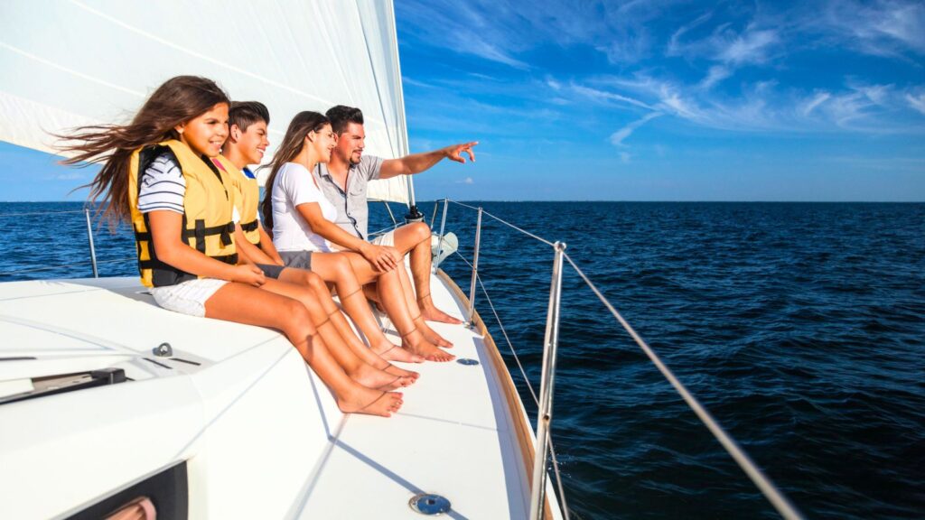 Four people sit on the deck of a sailboat, enjoying some Labor Day boating as they face the open ocean. One person points toward the horizon. All are barefoot, and the weather appears sunny with a clear blue sky.