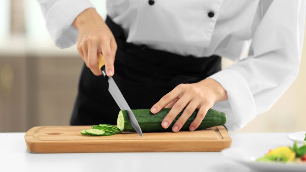 A person in a chef’s uniform, inspired by the World’s Best Female Chef, slices a cucumber on a wooden cutting board with a knife—perhaps preparing ingredients for an innovative cucumber dessert.
