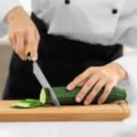 A person in a chef’s uniform, inspired by the World’s Best Female Chef, slices a cucumber on a wooden cutting board with a knife—perhaps preparing ingredients for an innovative cucumber dessert.