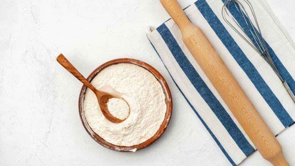 A wooden bowl of flour with a spoon next to a blue and white striped towel, rolling pin, and metal whisk on a white surface.