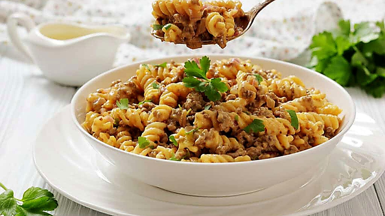 A white bowl of Cheeseburger Hamburger Helper, garnished with parsley, sits on a white plate, with a fork holding a bite above the bowl.