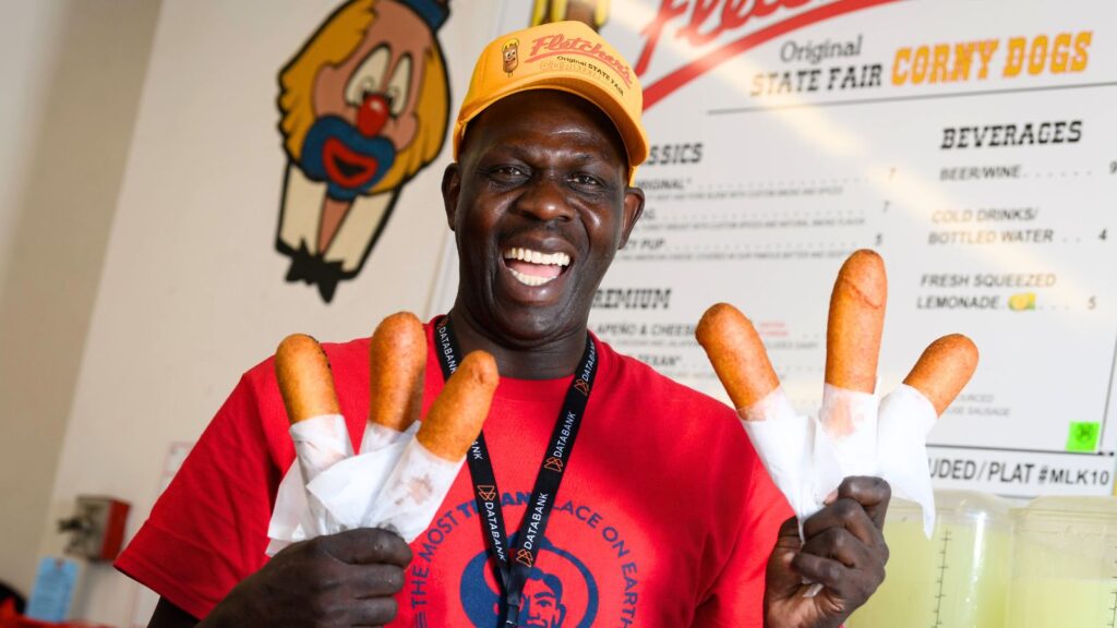 Man in a red shirt and yellow hat smiles while holding six corn dogs in front of a concession stand menu and cartoon logo.
