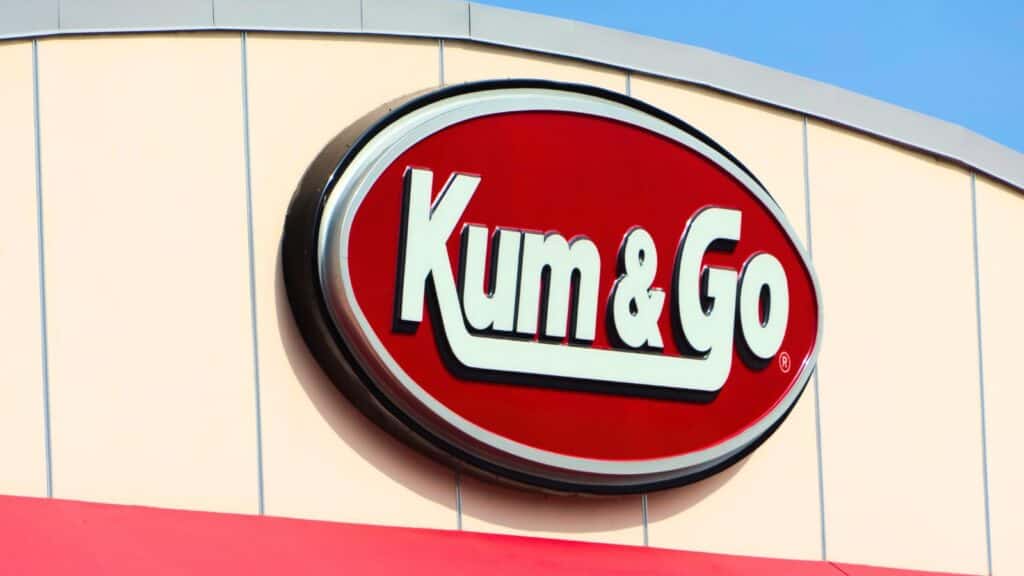 Red and white Kum & Go convenience store sign on a building exterior, shown in bright daylight against a pale wall with a blue sky background.