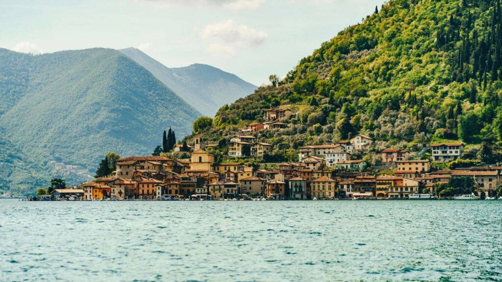 A small lakeside town with colorful buildings sits at the base of green, forested hills and mountains, under a partly cloudy sky.