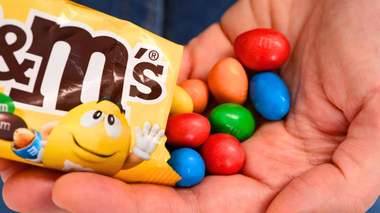 A hand holds a handful of colorful peanut M&M’s candies next to an open yellow M&M’s package.