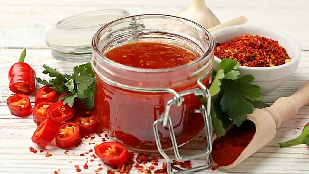 A jar of red chili sauce on a wooden surface, surrounded by sliced red chilies, parsley, garlic, chili flakes, and a wooden scoop with ground chili powder.