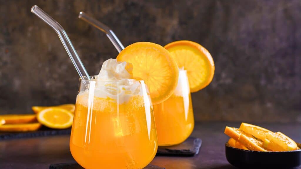 Two glasses of orange soda with whipped cream, glass straws, and orange slices; bowl of orange wedges on the side.