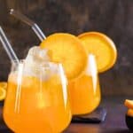 Two glasses of orange soda with whipped cream, glass straws, and orange slices; bowl of orange wedges on the side.