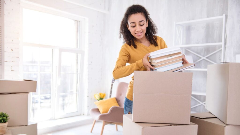 A woman packs a stack of books into a cardboard box in a bright room with moving boxes, shelves, and a chair visible in the background, preparing for college move-in day.