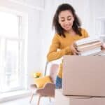 A woman packs a stack of books into a cardboard box in a bright room with moving boxes, shelves, and a chair visible in the background, preparing for college move-in day.