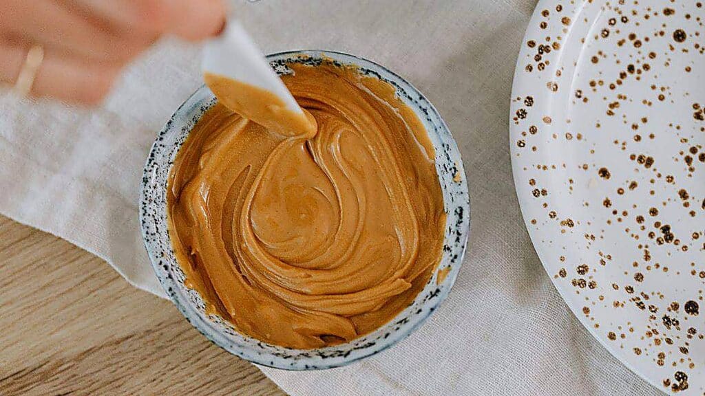 A small ceramic bowl filled with creamy peanut butter sits on a table next to a patterned plate, with a hand holding a white spatula in the bowl.