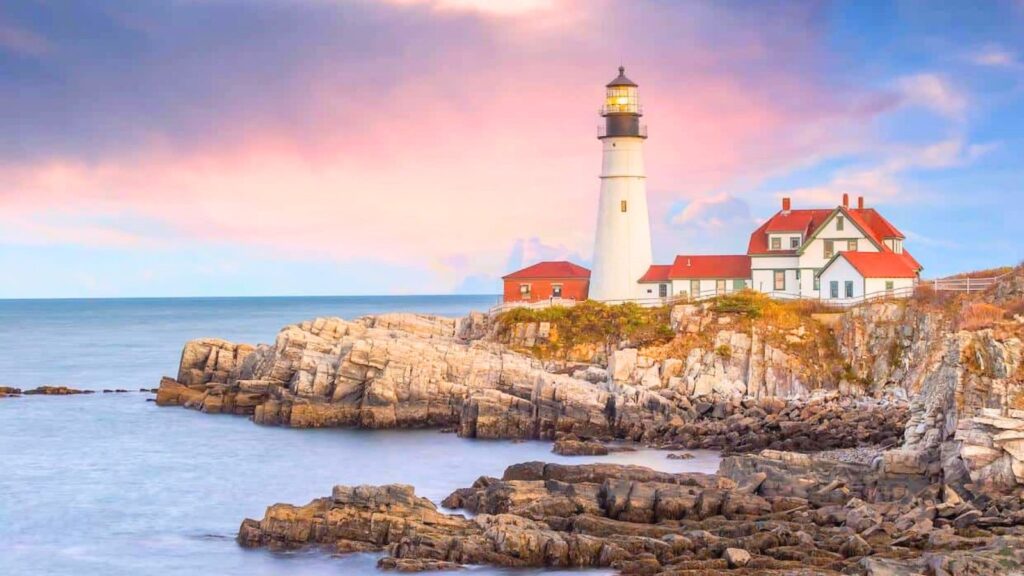 A white lighthouse with red-roofed buildings sits on a rocky coastline by the ocean under a pink and blue sky.