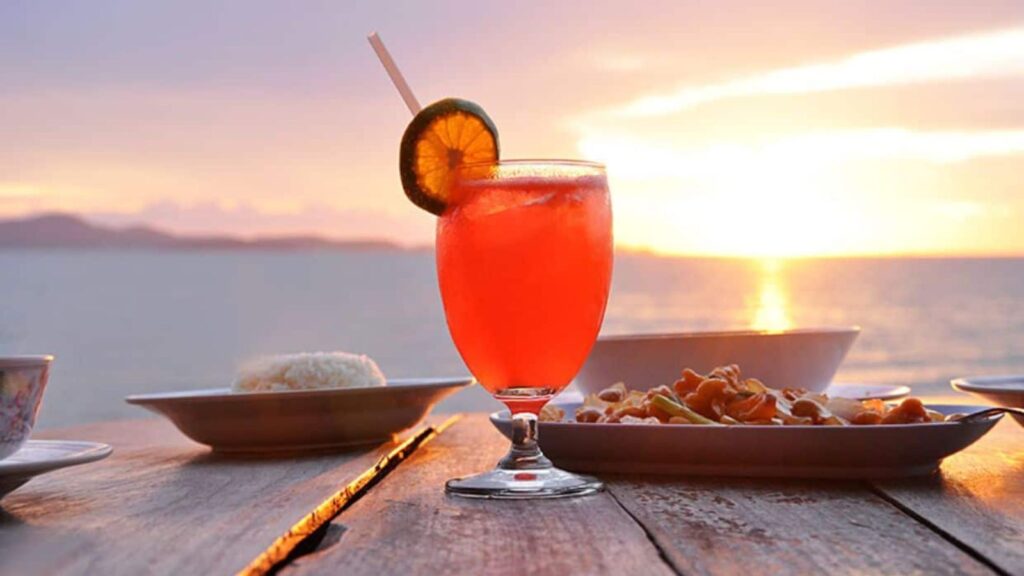 A red cocktail with a lime slice and straw sits on a wooden table with plates of food, overlooking the ocean at sunset.
