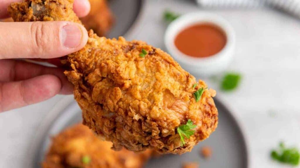A hand holding a piece of crispy fried chicken drumstick, with sauce and more chicken on a plate in the background.