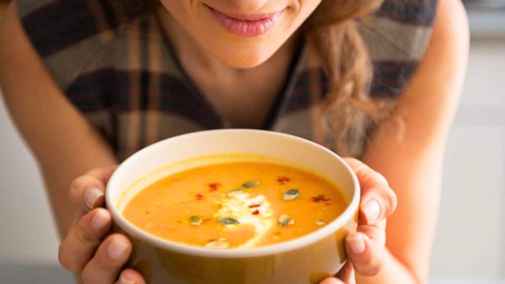 Woman holding a bowl of creamy orange soup topped with seeds and a swirl of cream, smiling slightly.