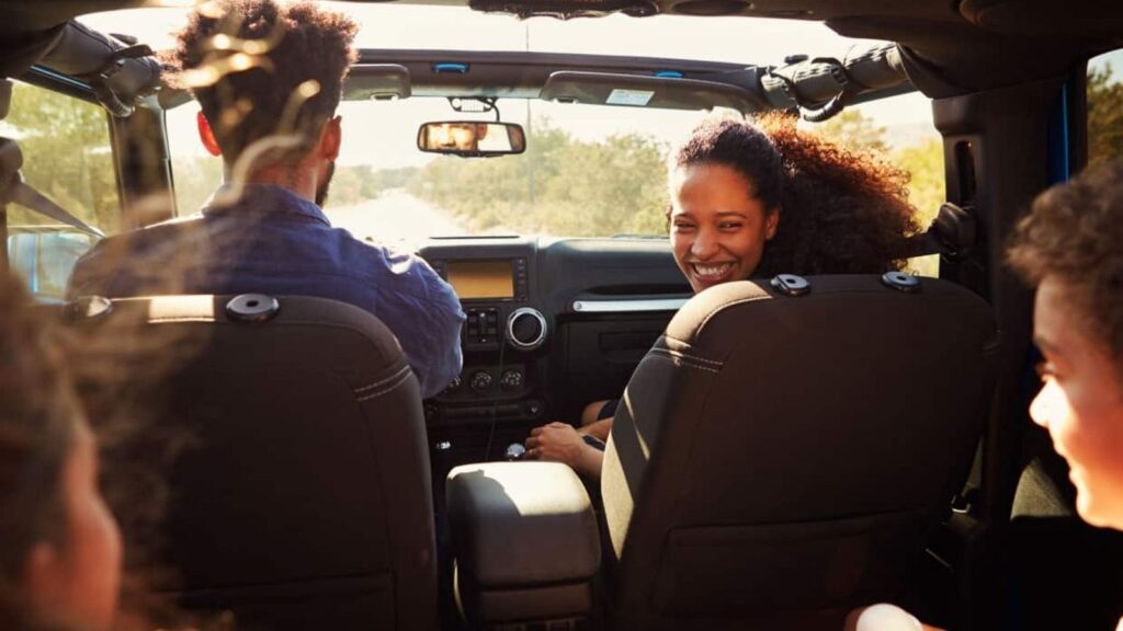 A man drives a convertible with three passengers, including a woman smiling in the front. Trees and sunlight are visible outside.