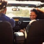 A man drives a convertible with three passengers, including a woman smiling in the front. Trees and sunlight are visible outside.