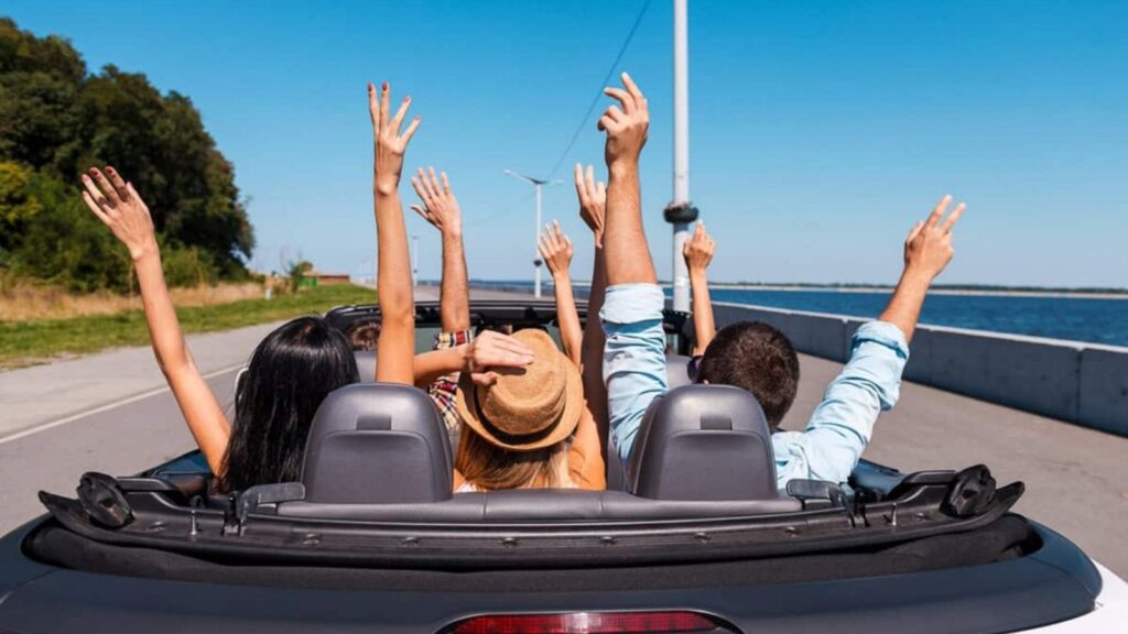 Four people in a convertible car with the top down, driving on a sunny road by the water, raising their arms in the air.