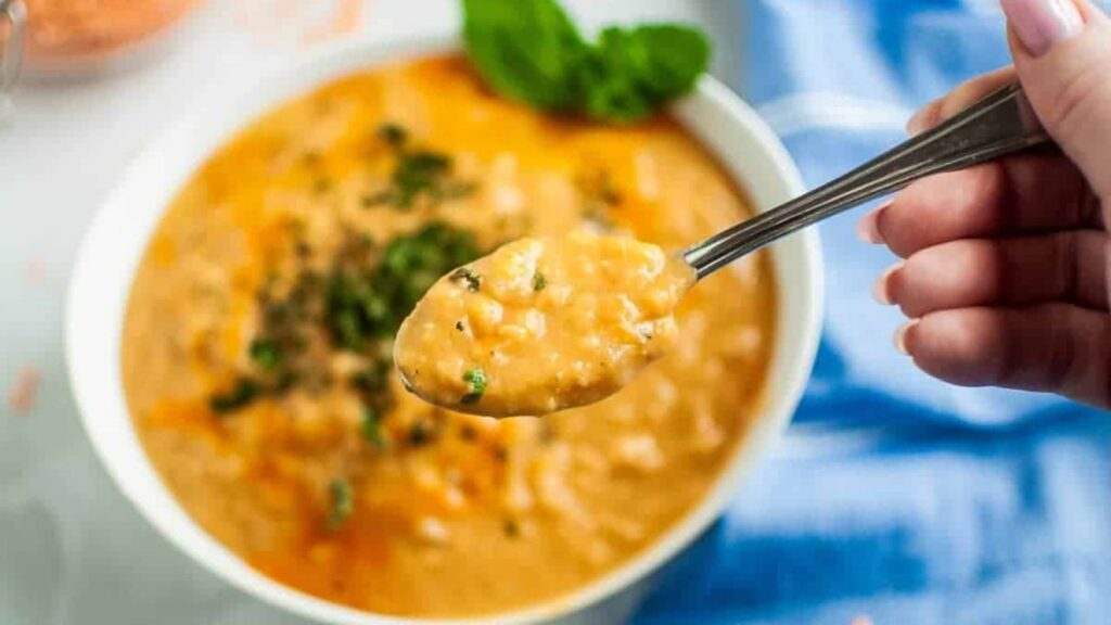 A hand holds a spoonful of creamy orange soup with herbs above a bowl of the same soup, with a blue cloth nearby.